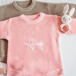 Puede incluir: Dos su&eacute;teres de beb&eacute; de punto, uno en color marr&oacute;n claro y otro en rosa suave. El su&eacute;ter rosa tiene el nombre "Amy" bordado en blanco. Tambi&eacute;n se ve un juguete de conejito de ganchillo blanco con un anillo de madera.