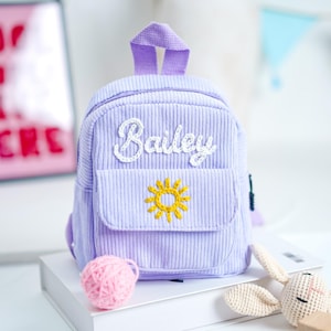 May include: A small, lavender corduroy backpack with the name "Bailey" embroidered in white script. A yellow sun design is embroidered on the front pocket. The backpack has a top handle and a small front pocket.