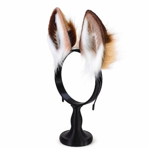 May include: A black headband featuring two upright, pointed animal ears made of faux fur. The ears display a gradient of colors, transitioning from dark brown at the tips and outer edges to a lighter tan, with long, soft white fur lining the inner ear.
