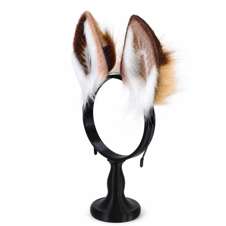 May include: A black headband featuring two upright, pointed animal ears made of faux fur. The ears display a gradient of colors, transitioning from dark brown at the tips and outer edges to a lighter tan, with long, soft white fur lining the inner ear.
