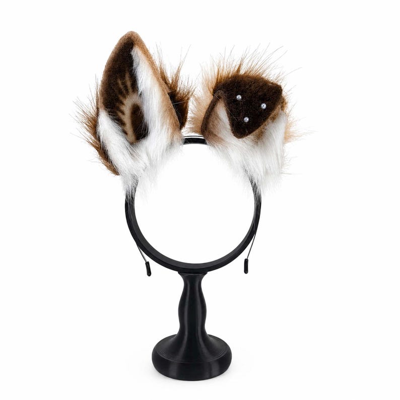 May include: A headband featuring two faux fur animal ears. The left ear is tall and pointed with brown, white, and black fur. The right ear is folded downward, featuring brown fur with three small white pearl-like accents on the surface.