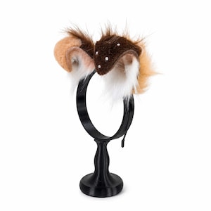May include: A headband featuring two plush, faux fur animal ears in shades of brown, tan, and white. The dark brown ear is accented with small, white spherical beads. The headband is displayed on a black, circular stand with a pedestal base.