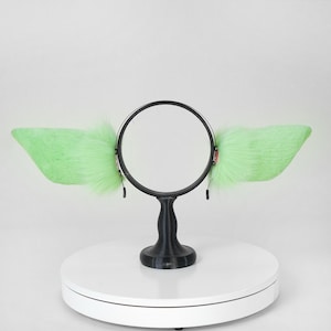 May include: A headband with large, light green, fluffy, faux fur ears. The ears are attached to a black circular frame with a black stand. The ears are a bright, vibrant green color.