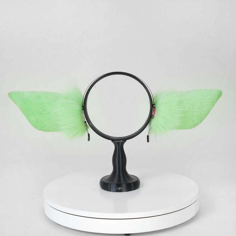 May include: A headband with large, light green, fluffy, faux fur ears. The ears are attached to a black circular frame with a black stand. The ears are a bright, vibrant green color.