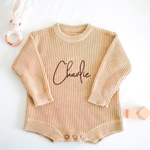 Puede incluir: Un body de beb&eacute; tejido de color beige con el nombre "Charlie" bordado en escritura marr&oacute;n. El body tiene mangas largas y cierres de bot&oacute;n. Un sonajero de conejito de ganchillo y bloques de madera tambi&eacute;n est&aacute;n en la imagen.