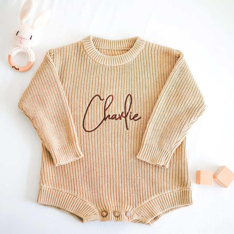 Puede incluir: Un body de beb&eacute; tejido de color beige con el nombre "Charlie" bordado en escritura marr&oacute;n. El body tiene mangas largas y cierres de bot&oacute;n. Un sonajero de conejito de ganchillo y bloques de madera tambi&eacute;n est&aacute;n en la imagen.