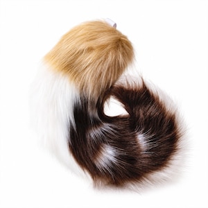 May include: A plush faux fur tail accessory featuring a tri-color pattern of tan, white, and dark brown. The long, soft synthetic fibers are arranged in a curved, curled shape, with a small white fabric loop attached at the top for secure fastening.