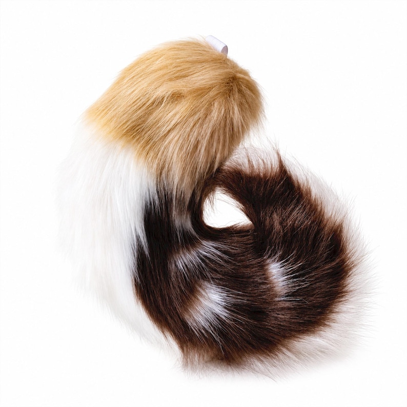 May include: A plush faux fur tail accessory featuring a tri-color pattern of tan, white, and dark brown. The long, soft synthetic fibers are arranged in a curved, curled shape, with a small white fabric loop attached at the top for secure fastening.