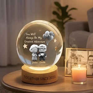 May include: A clear glass globe with a wooden base, featuring a couple figurine and the text "You Will Always Be My Greatest Adventure." The base reads "TOGETHER SINCE 2015." The globe is illuminated, with a photo frame and candle nearby.