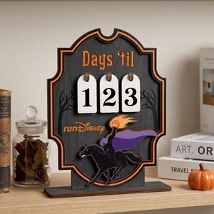 May include: A black and orange wooden countdown sign featuring the text Days 'til and runDisney. The sign includes three white hanging number tiles displaying 123 and a graphic of a headless horseman figure with a purple cape and orange pumpkin.