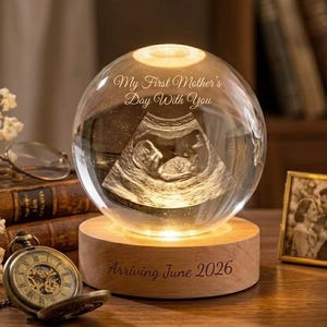 Op de afbeelding: Een heldere glazen bol met een echo-afbeelding van een baby, verlicht van binnen. De tekst "My First Mother's Day With You" en "Arriving June 2026" zijn gegraveerd. De bol staat op een houten voet.
