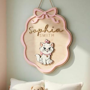 Könnte beinhalten: Ein dekoratives Wandschild aus Holz mit heller Holzoberfläche, rosa Rand und einer rosa Schleife oben. Das Schild zeigt den Namen Sophia in Schreibschrift, den Namen SMITH in Blockbuchstaben und eine Illustration eines weißen Kätzchens.