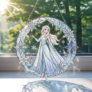 Può includere: Un'applique per finestra in vetro colorato raffigurante una principessa in un abito blu fluente, circondata da fiocchi di neve e una cornice circolare. L'opera d'arte è nei toni del blu e del bianco, con una catena per appenderla. La finestra è esposta davanti a una finestra.
