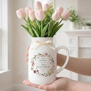 Könnte beinhalten: Eine weiße Keramikvase mit Henkel, gefüllt mit rosa Tulpen und Grün. Die Vase hat ein Blumengirlanden-Design und den Text "First mom EST. 1996 Now grandma EST. 2026". Ein cremefarbenes Band ist um die Vase gebunden.