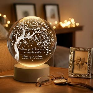 Puede incluir: Una esfera de cristal iluminada con un diseño de árbol y las palabras "Always & Forever in our hearts". La base es de color marrón claro. Una foto enmarcada, gafas y un reloj también están sobre la mesa.