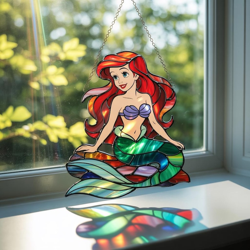 K&ouml;nnte beinhalten: Ein Buntglas-Suncatcher, der eine Meerjungfrau mit langem, flie&szlig;endem rotem Haar, einem violetten Muschel-Oberteil und einem gr&uuml;n-blauen Schwanz darstellt. Das St&uuml;ck h&auml;ngt an einer Metallkette und wirft eine bunte, durchscheinende Reflexion.
