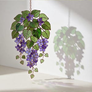Pu&ograve; includere: Decorazione floreale in vetro colorato con fiori viola e foglie verdi. L'opera d'arte &egrave; appesa a una catena. Il pezzo proietta un'ombra sul muro, evidenziando il motivo floreale. Un pezzo decorativo.
