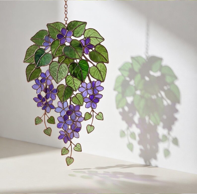 Pu&ograve; includere: Decorazione floreale in vetro colorato con fiori viola e foglie verdi. L'opera d'arte &egrave; appesa a una catena. Il pezzo proietta un'ombra sul muro, evidenziando il motivo floreale. Un pezzo decorativo.