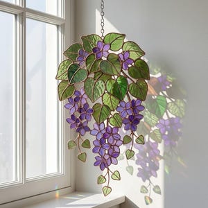 Pu&ograve; includere: Opera d'arte floreale in vetro colorato con fiori viola e foglie verdi. L'arte decorativa sospesa &egrave; sospesa da una catena ed &egrave; esposta davanti a una finestra. Il pezzo ha un bordo color rame.