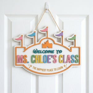May include: A white wooden wall sign shaped like a castle with orange trim and four colorful flags. The text reads "WELCOME TO MS. CHLOE'S CLASS" in multi-colored letters, with "THE HAPPIEST PLACE TO LEARN" printed below, suspended by a tan twine cord.