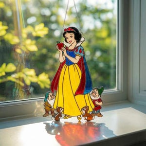K&ouml;nnte beinhalten: Ein Buntglas-Sonnenf&auml;nger mit einer Figur mit schwarzem Haar, blauem Oberteil und gelbem Rock, die einen roten Apfel h&auml;lt. Die Figur ist von zwei kleinen b&auml;rtigen Gestalten mit H&uuml;ten und zwei braunen Eichh&ouml;rnchen umgeben, an einer Kette h&auml;ngend.