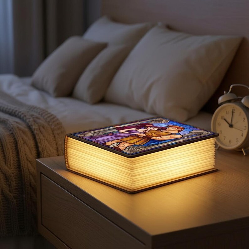 May include: A decorative book-shaped lamp with a stained-glass design on the cover, depicting a couple embracing. The lamp emits a warm, soft glow from the pages. It sits on a wooden nightstand next to a bed.