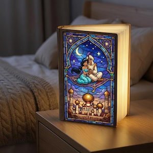 Może przedstawiać: Dekoracyjna lampa w kształcie książki z witrażowym wzorem przedstawiającym Aladyna i Jasminę na latającym dywanie. Lampa ma drewnianą ramę i emituje ciepłą poświatę, spoczywając na drewnianym stoliku nocnym. Grafika przedstawia pałac i gwiaździstą noc.