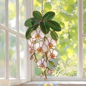 May include: A stained glass suncatcher featuring a cascading orchid plant with white petals, yellow centers, and deep green leaves. The piece is constructed with copper-toned metal soldering and hangs from a thin metal chain against a window.