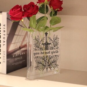 May include: Clear glass vase with a decorative sword and floral design, and the text "you do not yield." The vase holds several red roses and green leaves. A book with the words "Virtual Space" is visible in the background.