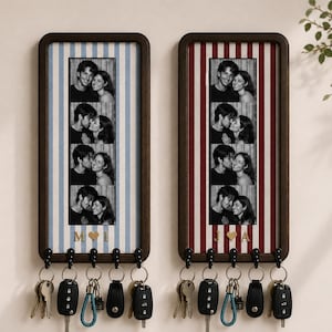 Op de afbeelding: Twee rechthoekige wand-sleutelhouders met donkerhouten lijsten. Elk is voorzien van een verticale fotostrip van een koppel en vijf zwarte haken met diverse sleutels. De ene heeft blauwe strepen met gouden letters M en L, de andere rode strepen met J en A.