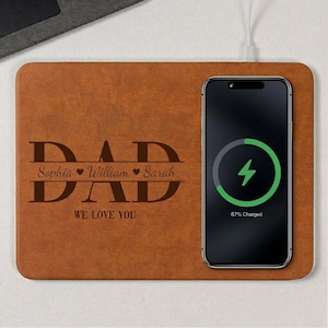 Puede incluir: Una base de carga inalámbrica de cuero marrón con la palabra "DAD" grabada en una fuente decorativa. Los nombres Sophia, William y Sarah están debajo de la palabra "DAD". Un teléfono inteligente negro se está cargando en la base.