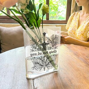 May include: A clear, rectangular vase with a black and white graphic of a sword and floral elements, with the text "you do not yield." The vase holds flowers and is set on a wooden table.