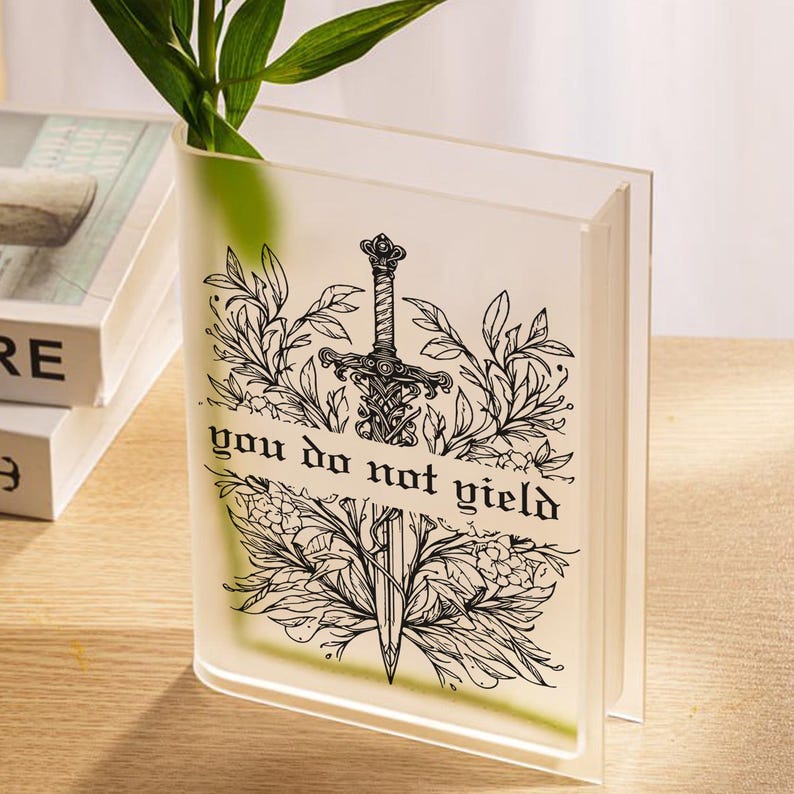 May include: A clear, book-shaped vase with a black illustration of a sword and foliage, and the text "you do not yield." Green leaves are visible inside the vase. The vase is made of clear acrylic.