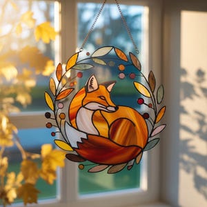 May include: Stained glass fox suncatcher with a copper chain. The fox is orange, white, and brown, curled up inside a wreath of leaves and berries. The leaves are shades of yellow, orange, and green.
