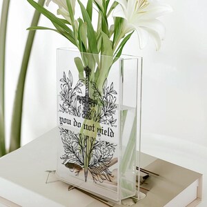 May include: Clear acrylic vase with a black and white illustration of a sword and floral elements. The vase has the text "you do not yield". White lilies and green foliage are inside the vase, which is sitting on a white surface.