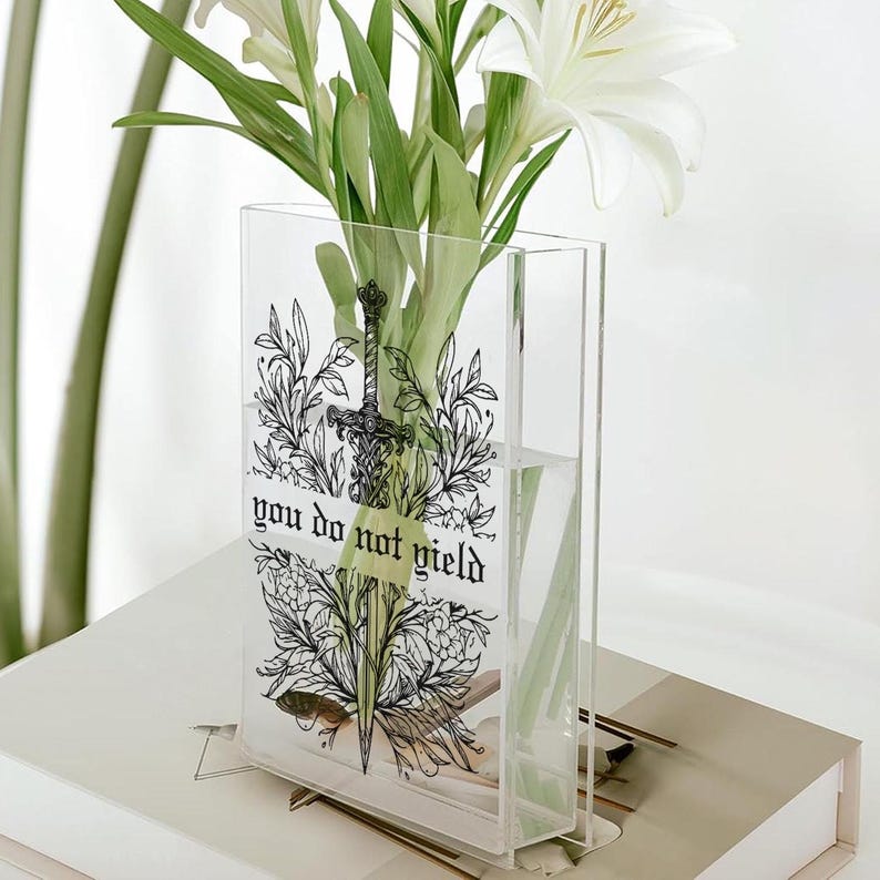 May include: Clear acrylic vase with a black and white illustration of a sword and floral elements. The vase has the text "you do not yield". White lilies and green foliage are inside the vase, which is sitting on a white surface.