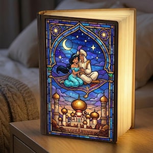 Puede incluir: Lámpara decorativa con forma de libro con un diseño de vidriera que representa a Aladdin y Jasmine en una alfombra mágica. La lámpara tiene un marco de madera y emite un brillo cálido. La vidriera muestra un cielo nocturno con estrellas, una luna creciente y un palacio.