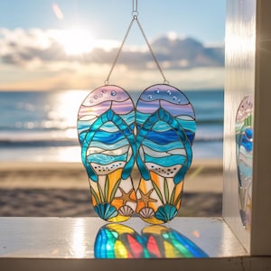 May include: Stained glass flip-flops with a beach scene design. The flip-flops feature blue, turquoise, and white waves, starfish, and seashells. The background shows a sunset over the ocean, creating a coastal theme.