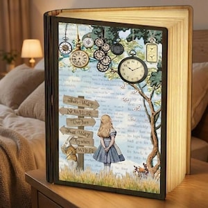 Puede incluir: Lámpara decorativa con forma de libro que presenta una escena de Alicia en el País de las Maravillas. Muestra a Alicia, relojes y señales direccionales. Hecha de madera, emite una luz cálida. Mide unos 25 cm de alto.