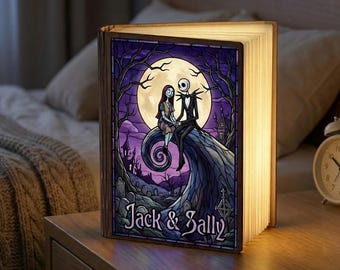 Puede incluir: Una lámpara de madera con forma de libro que presenta una ilustración de estilo vidriera de Jack y Sally de Pesadilla antes de Navidad. La obra de arte representa a los personajes sentados en una colina bajo la luna llena, con las palabras "Jack & Sally" debajo.