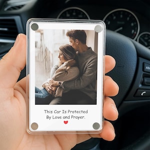 Könnte beinhalten: Ein klarer, rechteckiger Auto-Magnet mit einem Foto eines Paares, das sich umarmt. Der Magnet trägt den Text "This Car Is Protected By Love and Prayer" mit einem roten Herzen. Der Magnet wird in einer Hand gehalten.