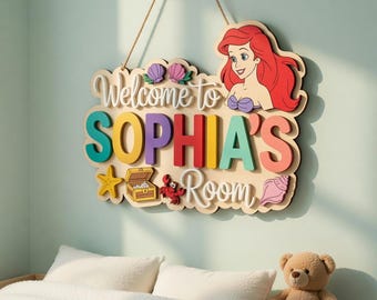 Può includere: Cartello in legno con la scritta "Welcome to SOPHIA'S Room" con un'illustrazione a cartoni animati di Ariel de La Sirenetta. Il cartello include una stella marina, un forziere e una conchiglia.