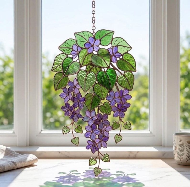 Pu&ograve; includere: Decorazione a sospensione in vetro colorato raffigurante fiori viola e foglie verdi. L'opera &egrave; sospesa a una catena. Lo sfondo mostra una finestra e la luce naturale. Un elemento decorativo.