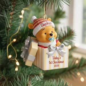 Puede incluir: Un adorno navideño de Winnie the Pooh. El adorno presenta a Winnie the Pooh con un gorro a rayas rojas y blancas y un chupete. Está dentro de una caja de regalo con el texto "Baby's 1st Christmas 2025". La caja tiene un lazo plateado.