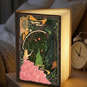 Puede incluir: Lámpara decorativa con forma de libro con una cubierta detallada que presenta a una princesa con un vestido rosa y una bruja verde. La cubierta tiene la palabra "Wicked" y un castillo verde. Las páginas iluminadas emiten un brillo cálido.