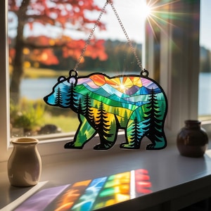 May include: A stained glass bear suncatcher with a colorful mountain and forest scene. The bear is outlined in black and hangs from a silver chain. The scene includes shades of blue, green, yellow, orange, and red.