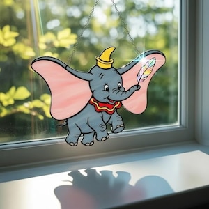 May include: A stained glass suncatcher depicting a gray elephant with large pink ears, a yellow hat, and a red and yellow collar, holding a multicolored feather in its trunk. The piece hangs by a silver chain against a window pane.