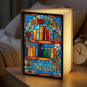May include: A decorative book-shaped lamp with a stained-glass design. The lamp features a colorful illustration of books on shelves, surrounded by flowers, with the name "Sarah" in cursive. The lamp is lit, with the pages glowing.