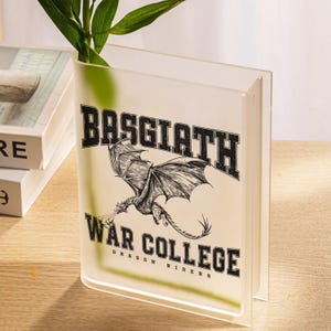 May include: Clear acrylic book-shaped vase with the text "BASGIATH WAR COLLEGE DRAGON RIDERS" and a dragon illustration. The vase holds green plant stems. The design is in black and white.