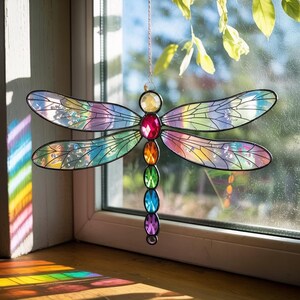 May include: A colorful stained glass dragonfly suncatcher. The dragonfly features iridescent wings and a body adorned with a series of jewel-toned gems. The suncatcher is suspended in front of a window, catching the light.
